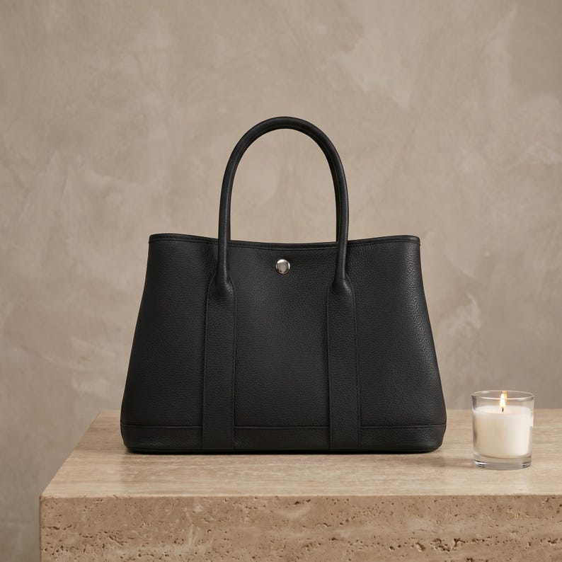 Black handmade leather tote bag shown front-on, structured horizontal cowhide shoulder bag with tall rolled handles, snap-stud top and silver-tone hardware.