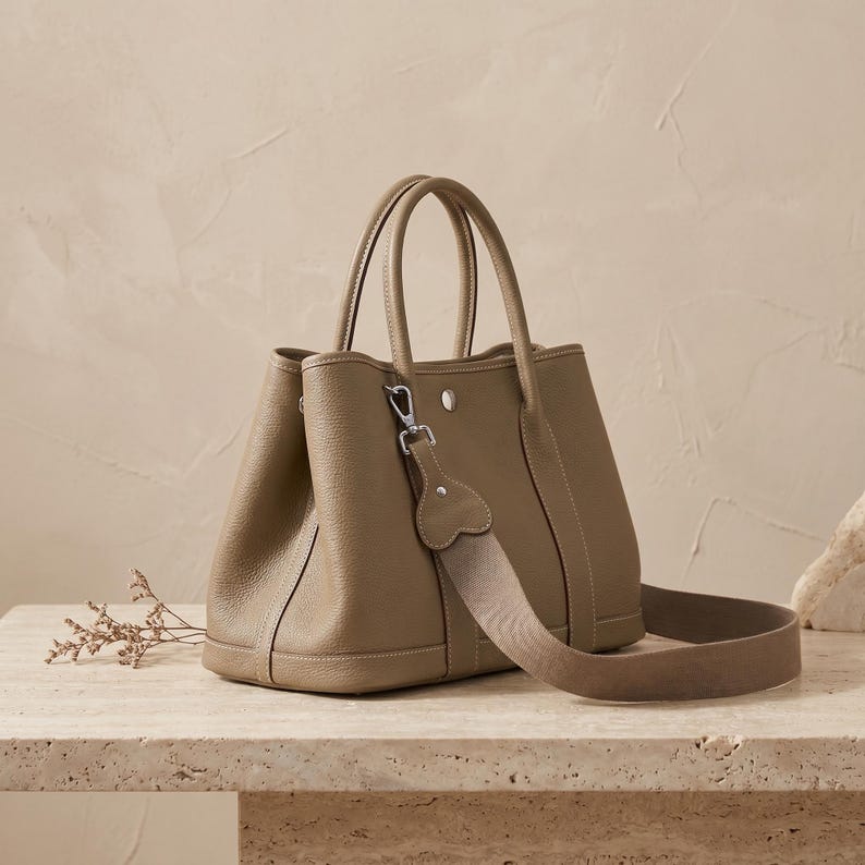 Khaki leather tote bag displayed with its removable adjustable webbing and leather crossbody strap and silver-tone clip hardware laid out beside the shoulder bag.