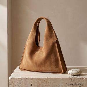 Pode incluir: Uma bolsa hobo de camurça castanha com silhueta triangular, apresentando uma alça única integrada e uma textura suave e mate. A bolsa tem costuras estruturadas ao longo das laterais e uma abertura superior. O texto ArcaLabLeather é visível.