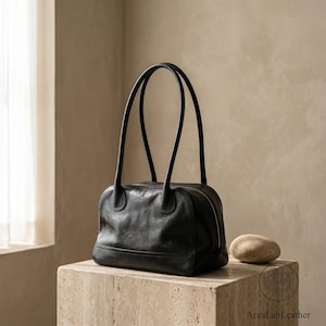 Puede incluir: Un bolso de mano de cuero negro con acabado liso y pulido, que presenta una base rectangular, esquinas superiores redondeadas y dos correas de hombro largas y delgadas. Una cremallera plateada recorre el borde superior, con la marca ArcaLabLeather.