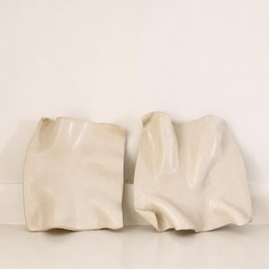 May include: Two off-white ceramic plates with irregular, wavy, and rippled edges. The surfaces have a smooth, matte finish with subtle variations in tone, creating a fluid, organic shape that resembles folded fabric or soft, undulating waves.