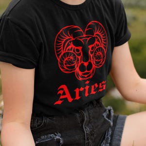 Könnte beinhalten: Ein schwarzes T-Shirt mit kurzen Ärmeln und einem leuchtend roten Grafikdruck eines Widderkopfes mit großen, geschwungenen Hörnern. Unter dem Widder-Motiv steht das Wort Aries in einer stilisierten roten Schrift. Rundhalsausschnitt und Ärmelaufschläge.