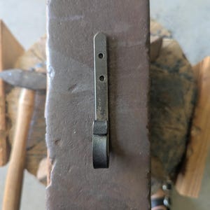 May include: A hand-forged metal hook with a flat, rectangular mounting plate featuring two circular pre-drilled holes. The hook has a dark, textured, matte finish and a curved, rounded end, resting on a heavy, rectangular metal anvil surface.