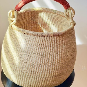 May include: A large, woven basket with a rounded shape and a red leather-wrapped handle. The basket is made of natural-coloured fibres and sits on a black surface. The basket's design is simple and functional.