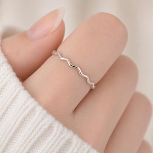 May include: A thin, polished silver ring featuring a continuous, delicate wavy or serpentine pattern that encircles the finger. The metal has a smooth, reflective surface, showcasing a simple, minimalist design that sits flush against the skin.