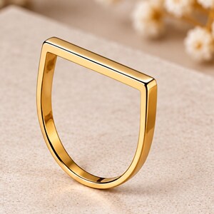 Könnte beinhalten: Ein polierter Goldring mit einem markanten geometrischen Design, bestehend aus einer flachen, horizontalen oberen Leiste, die mit einem geschwungenen, halbkreisförmigen Band verbunden ist. Die Metalloberfläche ist glatt und reflektierend.
