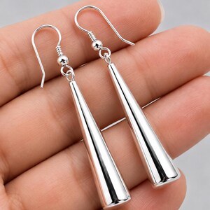 May include: A pair of silver-toned drop earrings featuring long, smooth, tapered conical pendants. Each earring hangs from a simple curved hook wire connected to a small spherical bead, creating a clean, polished, and reflective metallic finish.
