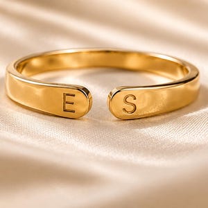 May include: A gold-toned open ring featuring a smooth, polished finish with the letter E engraved on one end and the letter S engraved on the other end. The band is adjustable, creating a gap between the two ends, and has a rounded, minimalist profile.