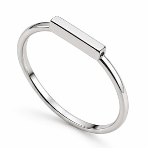 Peut inclure: Une bague en métal argenté poli composée d'un anneau fin et circulaire surmonté d'une barre rectangulaire horizontale. La surface métallique est lisse et réfléchissante, présentant une finition propre sur l'ensemble du bijou.