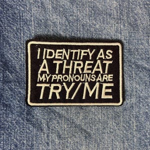 May include: A rectangular black embroidered patch with a white border and white stitched text that reads: I IDENTIFY AS A THREAT MY PRONOUNS ARE TRY/ME. The patch features a clean, bold font and is centered on a textured blue denim fabric background.