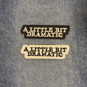 May include: Two rectangular embroidered patches featuring the text A LITTLE BIT DRAMATIC. One patch has a black background with white lettering, and the other has a cream background with black lettering. Both patches have a thick border.