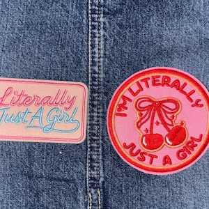 May include: Two embroidered patches on denim fabric. One rectangular patch is light pink with the text "Literally Just A Girl" in pink and blue script. The other is a circular pink patch featuring a red cherry graphic with a bow and the text "I'M LITERALLY JUST A GIRL" in red.