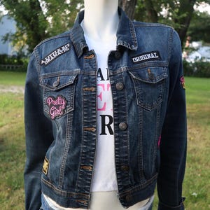 May include: A dark blue denim jacket featuring metal buttons and several embroidered patches, including text that reads A LITTLE BIT DRAMATIC, ORIGINAL, and Pretty Girl. The jacket is worn over a white shirt with partially visible black and pink text.