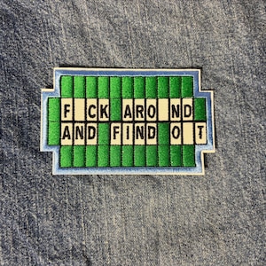 May include: A rectangular embroidered patch featuring a green grid design resembling a word puzzle game. The white letter tiles spell out the phrase FUCK AROUND AND FIND OUT in black text across two rows, surrounded by a light blue border.