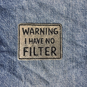 May include: A rectangular patch with a metallic silver background and a black border. The patch features the black embroidered text WARNING I HAVE NO FILTER in a bold, sans-serif font, centered on the surface of the patch.