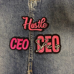 May include: Three embroidered patches featuring pink lettering with black borders. One patch reads Hustle in a cursive script, another reads CEO in bold block letters, and the third reads She is CEO with the words She is in white cursive script.