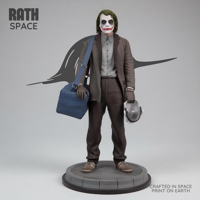 May include: A detailed statue of the Joker from The Dark Knight, featuring the character in a brown suit, gray shirt, and green hair. The figure holds a mask and a blue bag. The base reads "Crafted in Space Print on Earth."