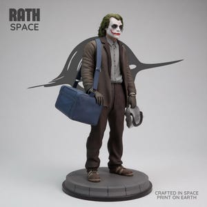 May include: A statue of the Joker character from the Batman franchise. The figure is wearing a brown suit, carrying a blue bag and a mask. The statue is on a gray circular base. The text "RATH SPACE" and "CRAFTED IN SPACE PRINT ON EARTH" are visible.