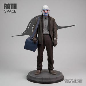 May include: A detailed statue of a figure in a clown mask, wearing a brown suit, gray shirt, and carrying a blue bag. The statue stands on a gray circular base. The text "RATH SPACE" and "CRAFTED IN SPACE PRINT ON EARTH" are visible.