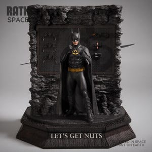 Puede incluir: Una estatua de Batman con un traje negro, cinturón amarillo y el símbolo del murciélago. La estatua está sobre una base oscura con el texto "LET'S GET NUTS". El fondo presenta una pared con batarangs y otros accesorios.
