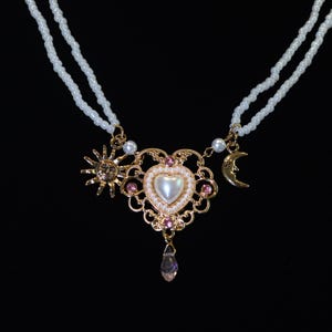 May include: A double-strand necklace featuring small white seed beads. The central pendant is a gold-toned filigree heart with a pearlescent center, pink accents, and a dangling crystal. A gold sun charm and a crescent moon charm are attached.