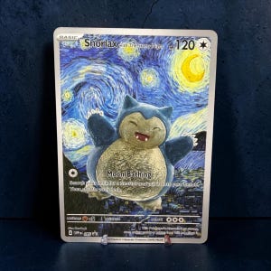 May include: A Snorlax with The Starry Night trading card featuring a blue and cream-colored Snorlax character centered against a swirling blue and yellow night sky background. The card displays the text Snorlax with The Starry Night, Moon Bathing, 120 HP, and various game statistics printed in black and white text across the card surface.