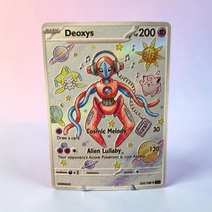 May include: A Deoxys trading card featuring a holographic finish with a hand-drawn illustration of the character wearing headphones. The card displays the text Cosmic Melody and Alien Lullaby, surrounded by colorful drawings of planets, stars, and a cassette player against a metallic silver background.