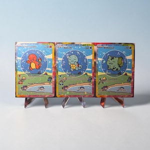 May include: A display of three trading cards featuring Charmander, Squirtle, and Bulbasaur. Each card has a unique illustration and text. The cards are displayed on clear acrylic stands, showcasing the artwork and design.