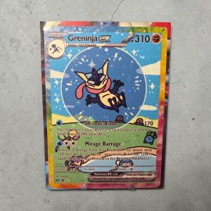 May include: A holographic Pokémon trading card featuring Greninja EX. The card has a blue and black color scheme with a central illustration of Greninja. Text includes "Shinobi Blade" and "Mirage Barrage".