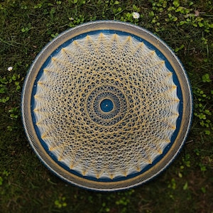 Op de afbeelding: Een cirkelvormig kunstwerk van spijkerkunst op een diepblauwe ondergrond, met een complex geometrisch patroon van witte en gouden draden gewikkeld rond kleine metalen spijkers in een radiaal ontwerp, wat een stervormig mandala-patroon vormt.