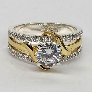 May include: A sterling silver ring with an S925 hallmark, featuring a central round-cut clear stone held by gold-toned prongs. The band consists of two outer rows of small clear stones and a smooth gold-toned center band that curves around the center.