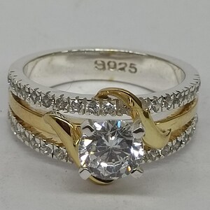May include: A two-tone ring featuring a central round-cut clear stone held by gold-colored prongs. The band consists of a silver-colored base with rows of small clear stones and a smooth gold-colored center inlay. The interior is stamped with 9925.