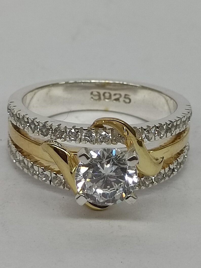May include: A two-tone ring featuring a central round-cut clear stone held by gold-colored prongs. The band consists of a silver-colored base with rows of small clear stones and a smooth gold-colored center inlay. The interior is stamped with 9925.