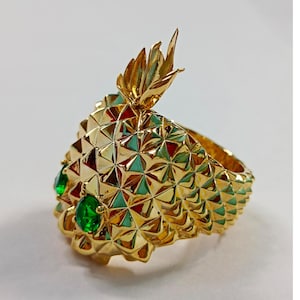 Op de afbeelding: Een goudkleurige ring in de vorm van een ananas. De ring heeft een gestructureerd oppervlak met een geometrisch patroon en groene edelsteenaccenten. De kroon van de ananas is gedetailleerd met goudkleurige bladeren.