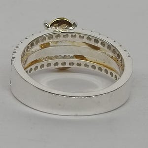 May include: A silver-toned ring featuring a wide band with a row of small, clear, circular stones embedded along the top edge. The interior of the ring displays a gold-toned accent, and a single, larger, round, light-brown stone is set at the top.