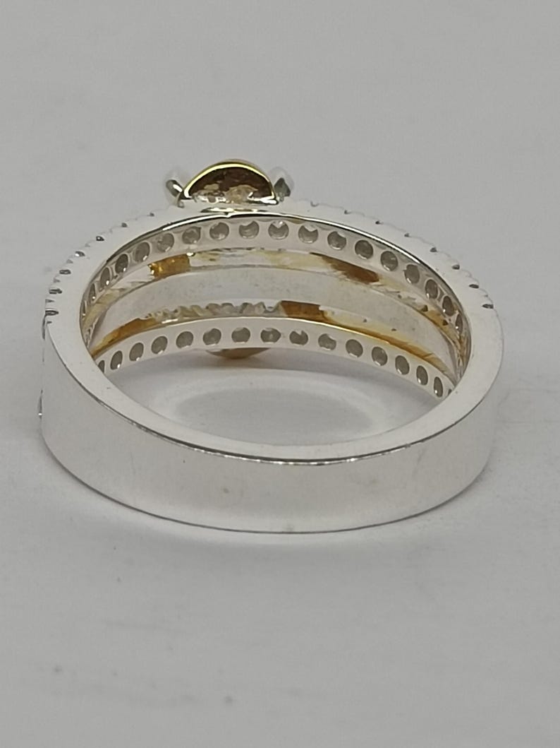 May include: A silver-toned ring featuring a wide band with a row of small, clear, circular stones embedded along the top edge. The interior of the ring displays a gold-toned accent, and a single, larger, round, light-brown stone is set at the top.