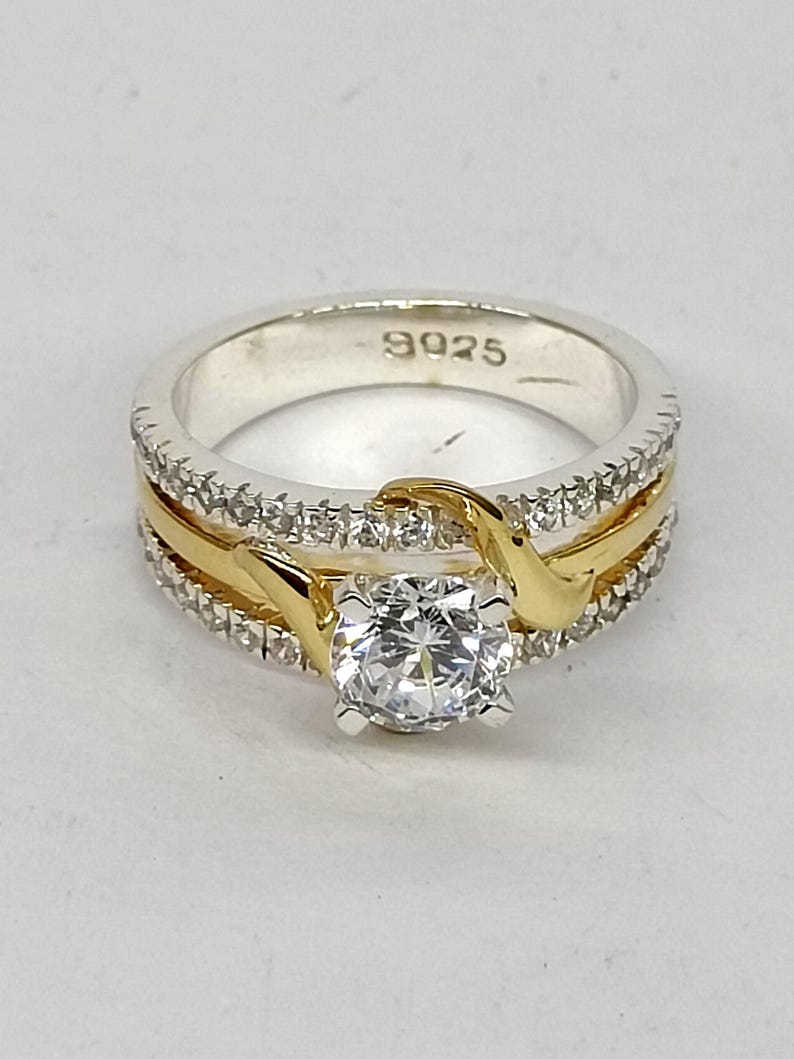 May include: A silver-toned ring featuring a central round-cut clear stone held by four prongs. The band includes a gold-toned curved accent and two rows of small clear stones. The interior of the band is clearly stamped with the text 9925.