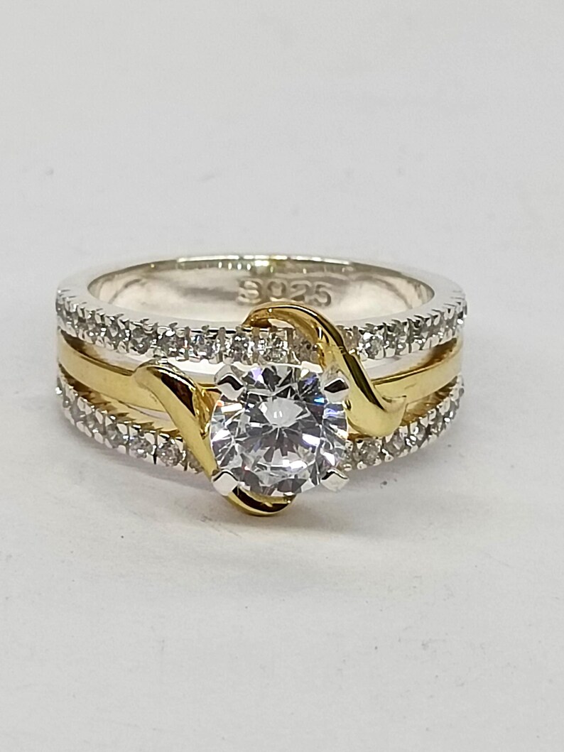 May include: A sterling silver ring with an S925 hallmark, featuring a central round-cut clear stone held by gold-toned prongs. The band consists of two outer rows of small clear stones and a smooth gold-toned center band that curves around the center.