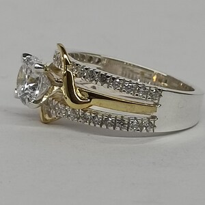 May include: A two-tone ring featuring a central round clear stone set in silver-colored metal. The band is composed of two silver-colored rows encrusted with small clear stones, separated by a smooth, curved gold-colored metal accent piece.