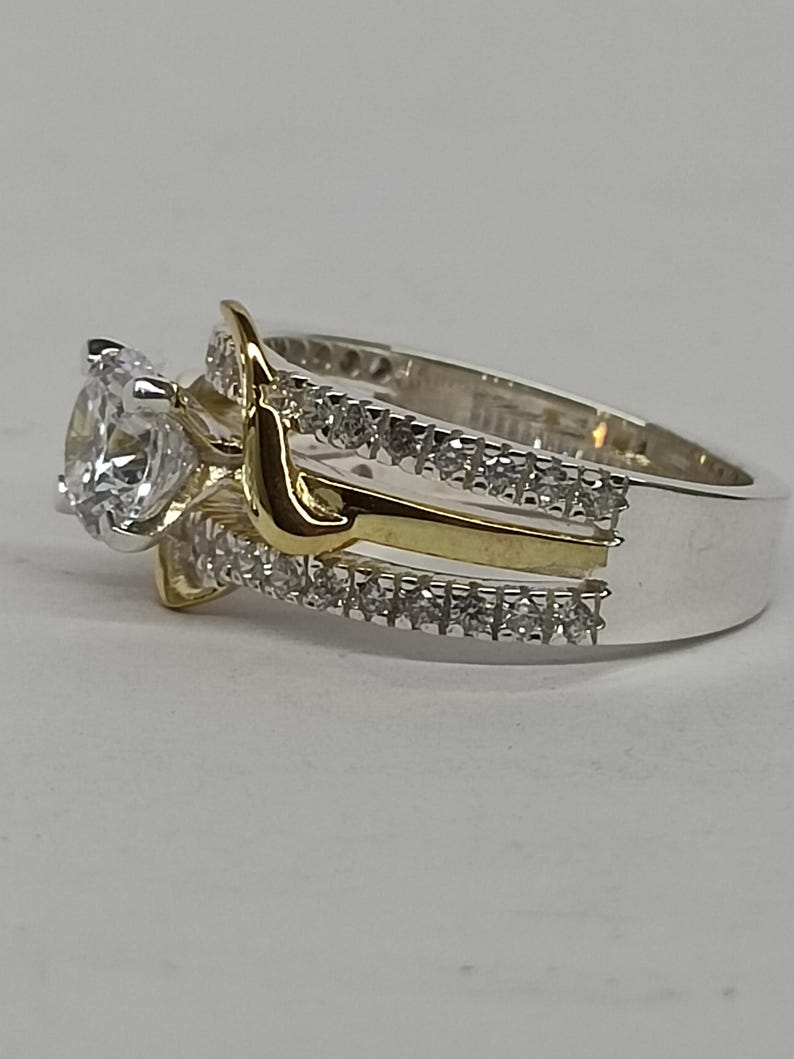 May include: A two-tone ring featuring a central round clear stone set in silver-colored metal. The band is composed of two silver-colored rows encrusted with small clear stones, separated by a smooth, curved gold-colored metal accent piece.