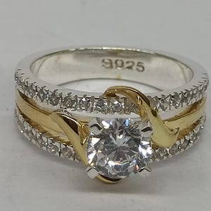 May include: A two-tone ring featuring a central round clear stone held by gold-colored prongs. The band consists of a central gold-colored strip flanked by two silver-colored bands encrusted with small clear stones. The inner band is stamped 925.