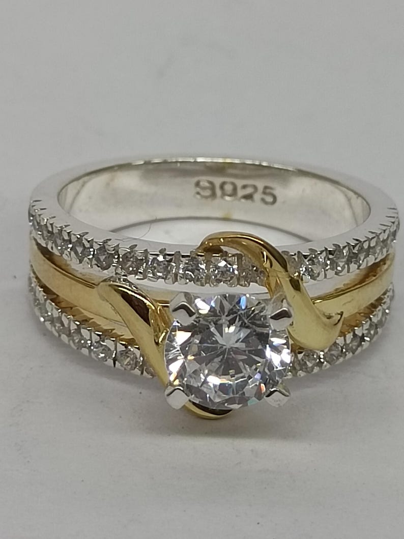 May include: A two-tone ring featuring a central round clear stone held by gold-colored prongs. The band consists of a central gold-colored strip flanked by two silver-colored bands encrusted with small clear stones. The inner band is stamped 925.