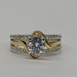 May include: A two-tone ring featuring a central round-cut clear stone held by a gold-colored bypass setting. The band consists of two outer rows of small pav&eacute;-set clear stones and a smooth gold-colored center band, creating a layered appearance.