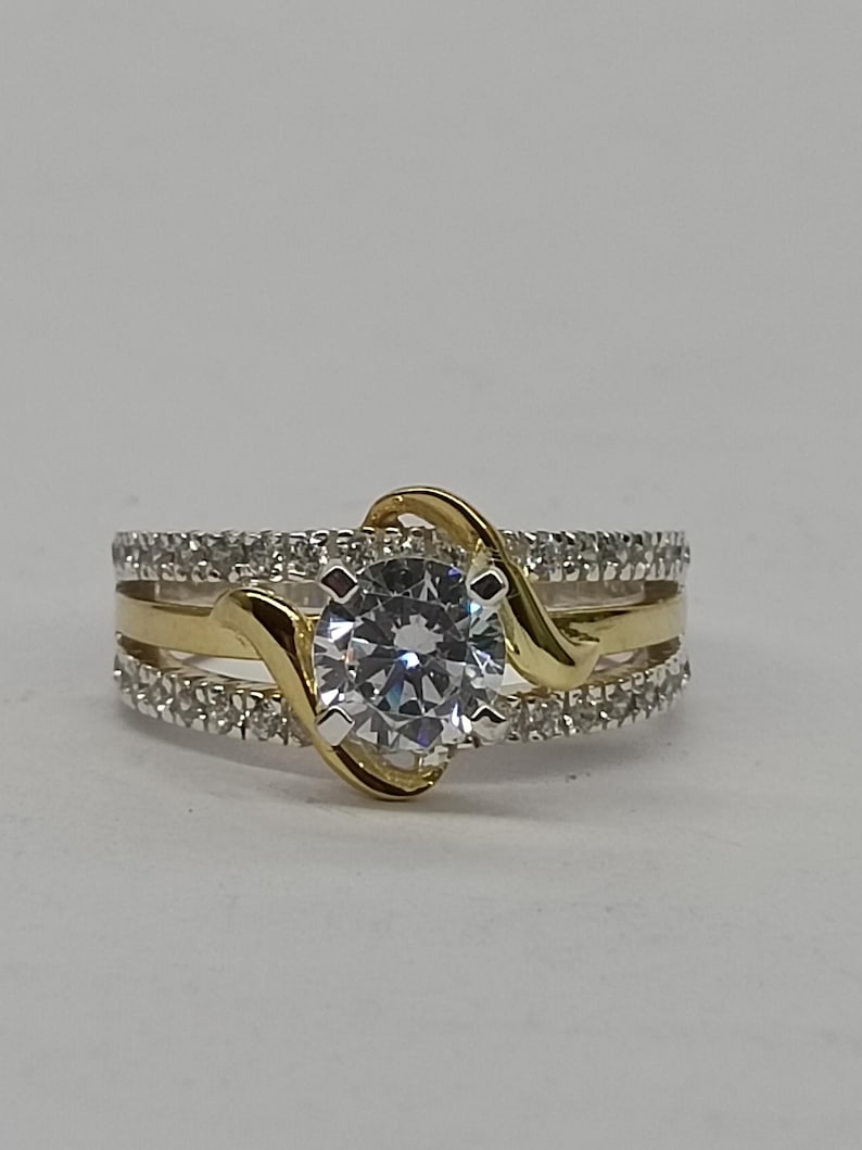 May include: A two-tone ring featuring a central round-cut clear stone held by a gold-colored bypass setting. The band consists of two outer rows of small pav&eacute;-set clear stones and a smooth gold-colored center band, creating a layered appearance.