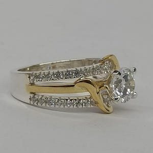 May include: A two-tone ring featuring a central round-cut clear stone held in a prong setting. The band is crafted from silver-colored metal with two rows of small clear stones and a central gold-colored metal accent that curves around the center.