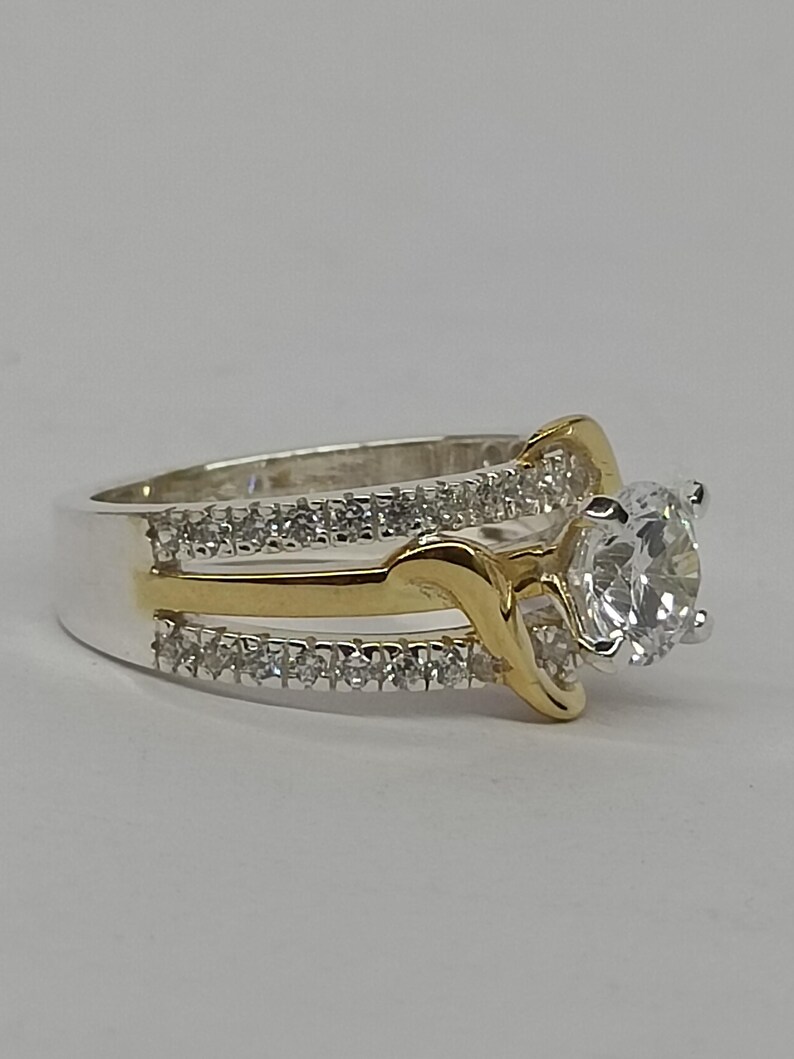 May include: A two-tone ring featuring a central round-cut clear stone held in a prong setting. The band is crafted from silver-colored metal with two rows of small clear stones and a central gold-colored metal accent that curves around the center.