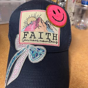 May include: A navy blue baseball cap featuring a rectangular patch with a mountain and sun illustration, the text "FAITH Can move mountains," a bright pink fuzzy smiley face patch, and a decorative blue and silver glittery bow applique.