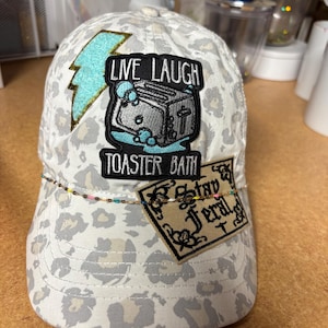 May include: A white baseball cap with a grey leopard print pattern. The cap features a glittery light blue lightning bolt patch, a black embroidered patch depicting a toaster in a bathtub with the text "LIVE LAUGH TOASTER BATH," and a tan rectangular patch with the text "Stay Feral." A thin, multi-colored beaded chain is draped across the front.