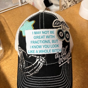 May include: A black and white trucker hat featuring a white rectangular patch with the text I MAY NOT BE GREAT WITH FRACTIONS, BUT I KNOW YOU LOOK LIKE A WHOLE BITCH. The hat includes a skeleton hand patch, a glittery teal star, and cherry patches.