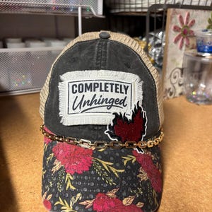 May include: A charcoal gray and tan mesh trucker hat featuring a rectangular patch with the text "COMPLETELY Unhinged" in black. The hat includes a gold-tone chain across the brim and a floral pattern with red flowers on the front panel.